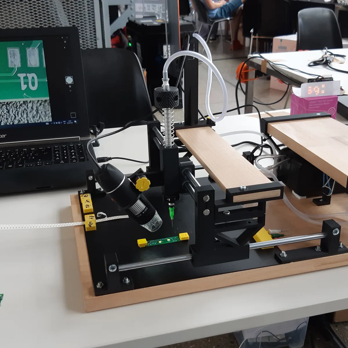 A DIY pick and place machine on a wooden base, with a vacuum nozzle, a USB microscope camera and a laptop showing the camera view